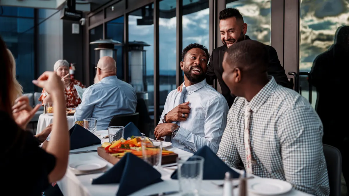 A group of people sit at a table with a white table cloth and a person wearing black greets two of the people.