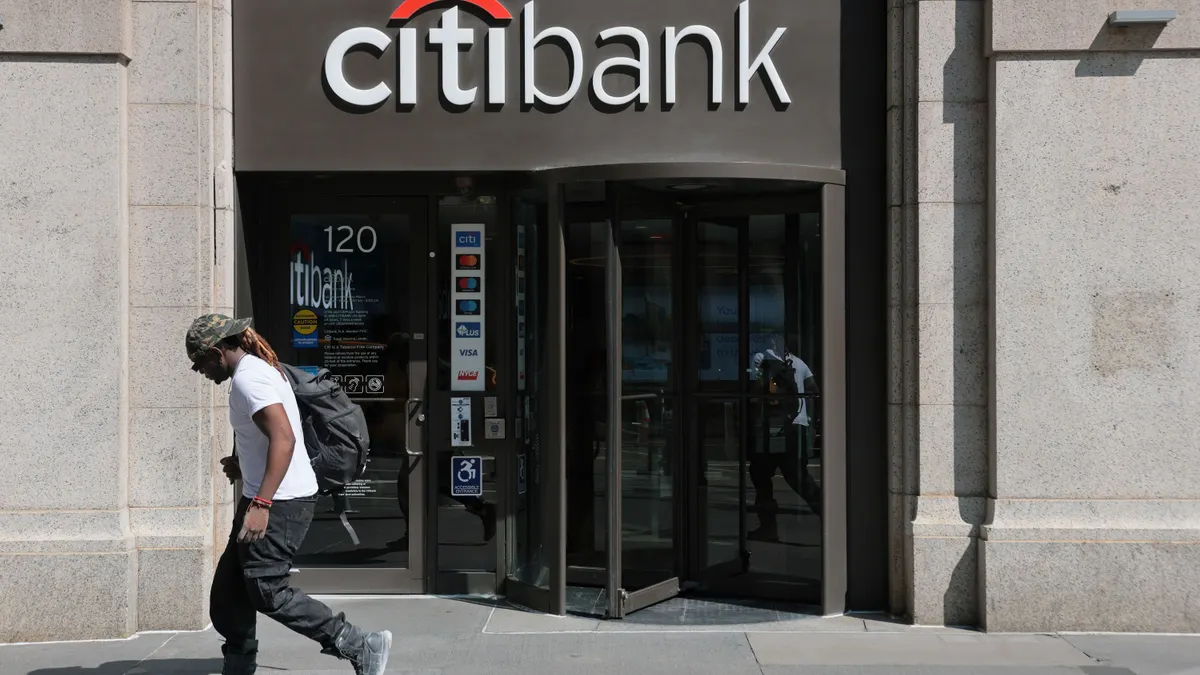A person walks past a building entrance with "Citibank" letters above the doorway.