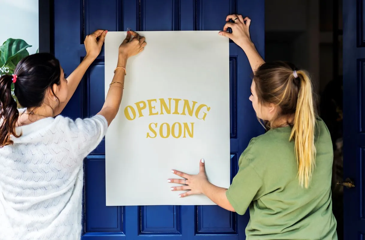 An image of two woment posting a white sign with yellow type that says &quot;opening soon&quot; on a blue door.