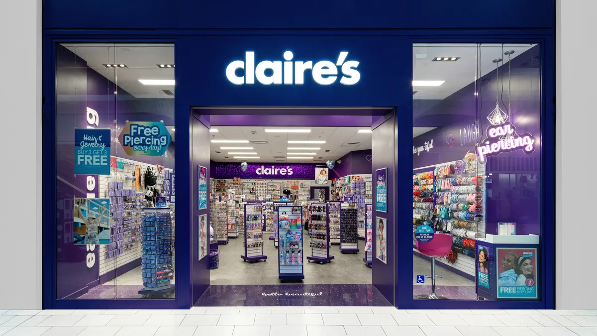 A glowing white "claire's" sign in lowercase block letters adorns a mall store trimmed in purple and dark blue. Windows signs advertise hair and jewelry specials and ear pierciing.