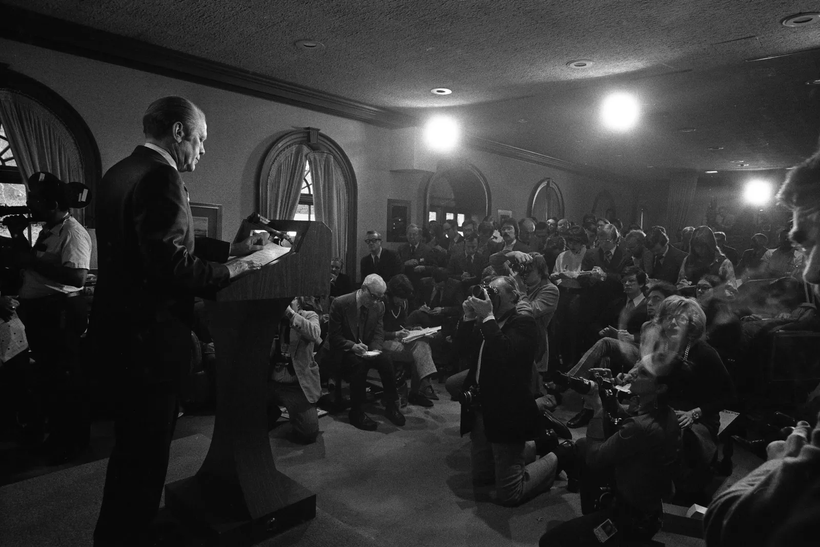 President Gerald Ford, left, stands at a podium in front of a crowded room of photographers and journalists at the White House.