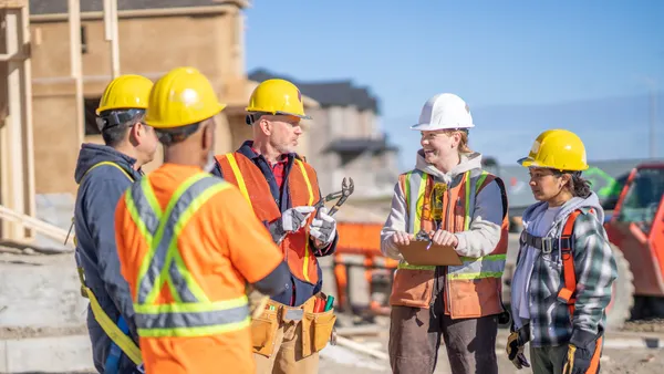 A small group of constructions workers meet with the forewoman to review the days work. They are each wearing proper safety equipment and are listening attentively.