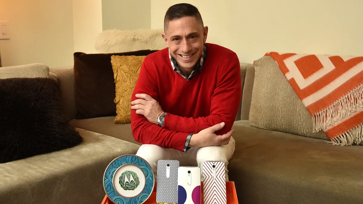 A person in a red sweater smiles at the camera while sitting on a beige couch. On a table in front of him are several phone cases and some housewares displayed on an orange tray.