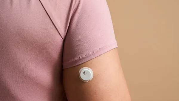 A round, white device is on a person's upper arm, below the sleeve of a pink shirt.