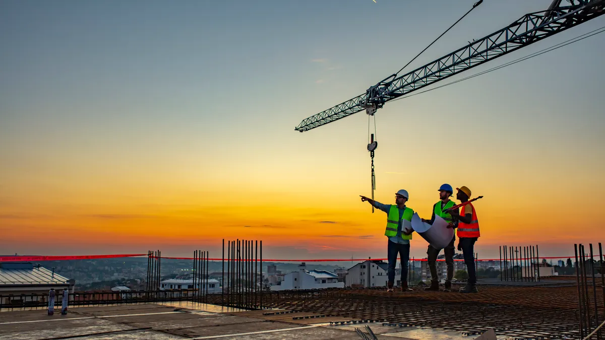 Three people in safety gear talk at the top of a building under construction.