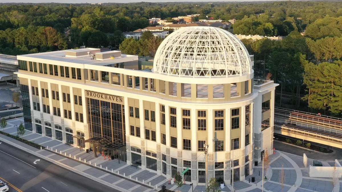 A large, newly constructed building looms above the treeline, with an open roof space and cars around.