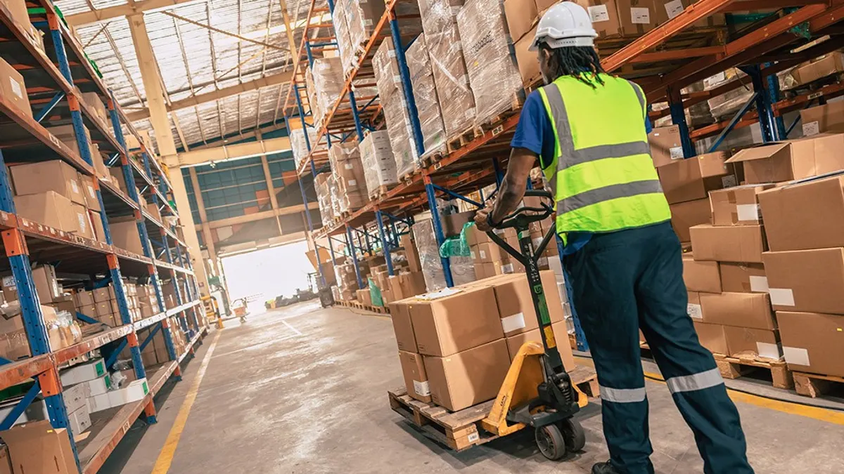 Warehouse worker in a hard hat and high-visibility vest moves a pallet of cardboard boxes with a manual pallet jack between tall storage racks.
