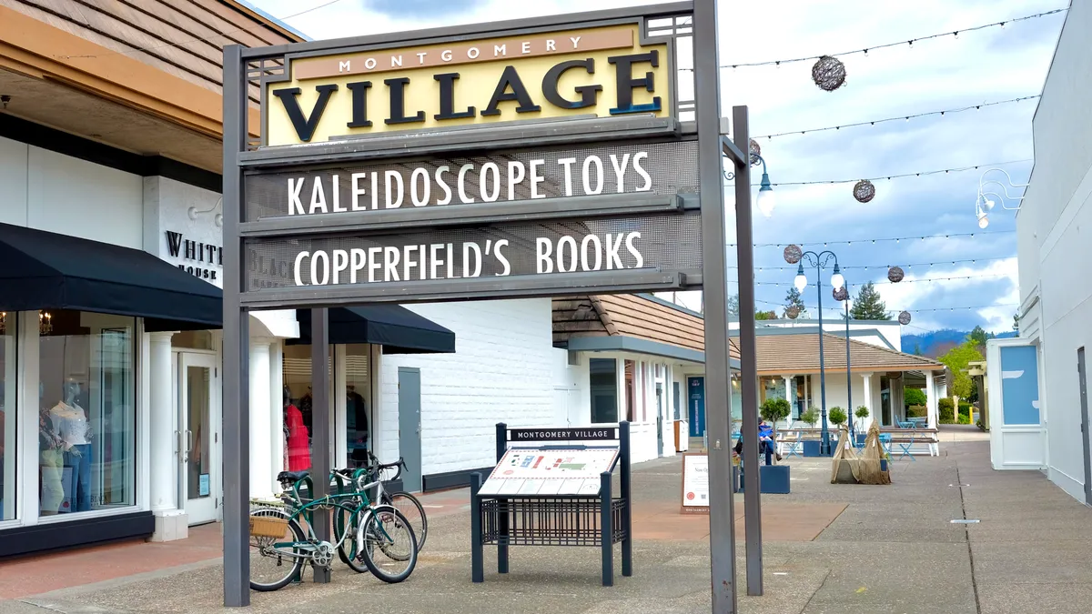 A large sign marks the entrance to an outdoor mall, with shops along a walkway, lights strung overhead and bikes parked to one side. A cloudy blue sky in the background.