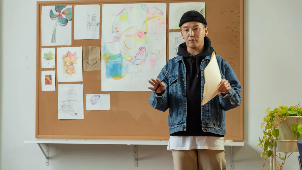 An Asian person stands in front of a whiteboard with art on it