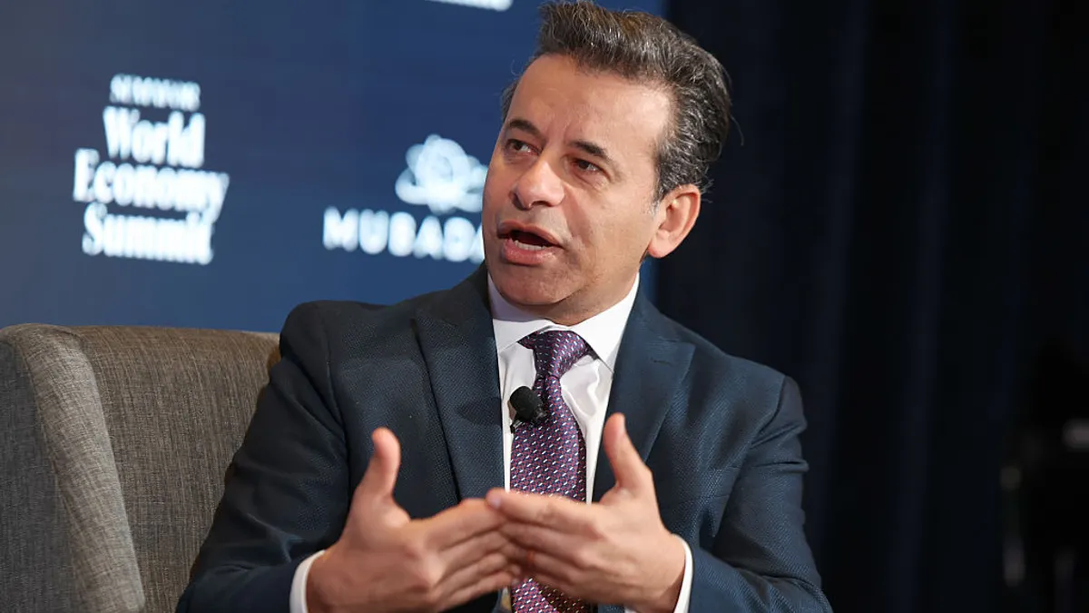 A person in a business suit gestures while speaking from a chair.