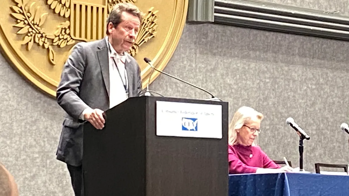 FDA Commissioner Robert Califf stands at a podium and speaks, while CFSAN Director Susan Mayne sits next to him at a table covered with a blue cloth.