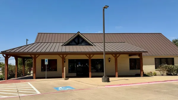 A beige brick building with a brown roof is shown, with a parking lot in front.