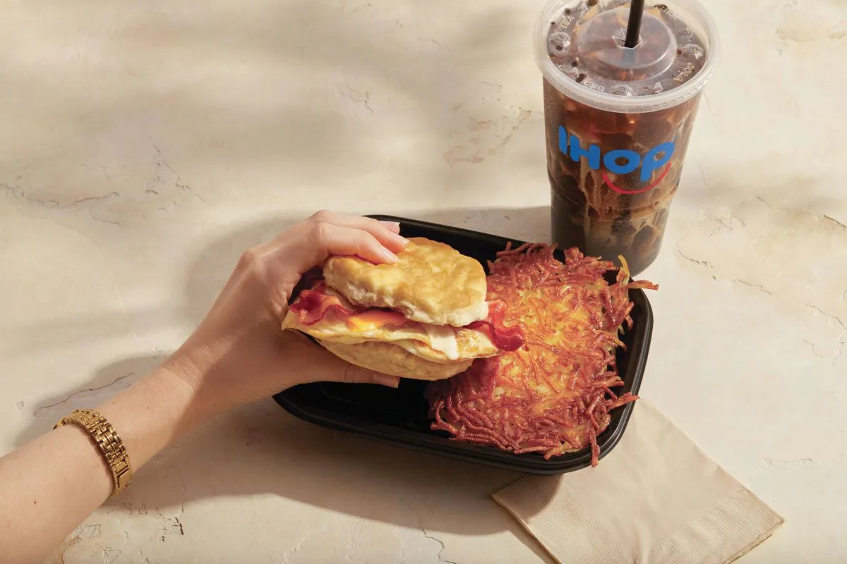 A woman's hand is holding a breakfast biscuit in a black takeout container with hashbrowns from IHOP.