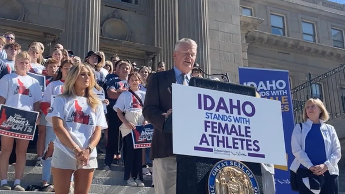 Brad Little stands behind a podium, flanked by advocates holding placcards