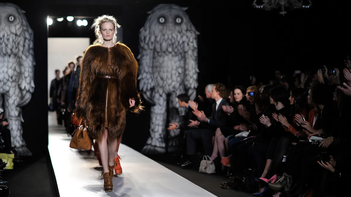 A model in a furry coat walks down a runway flanked by two large, stylized cartoon monsters.