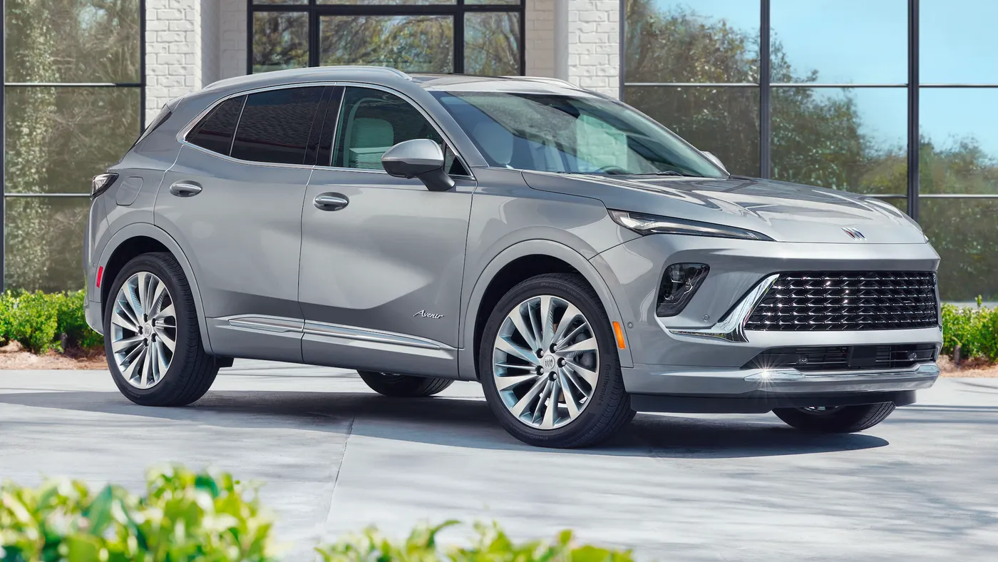 A front 3/4 view of the 2024 Buick Envision Avenir in Moonstone Gray Metallic paint parked in front of a building with greenery in the foreground.