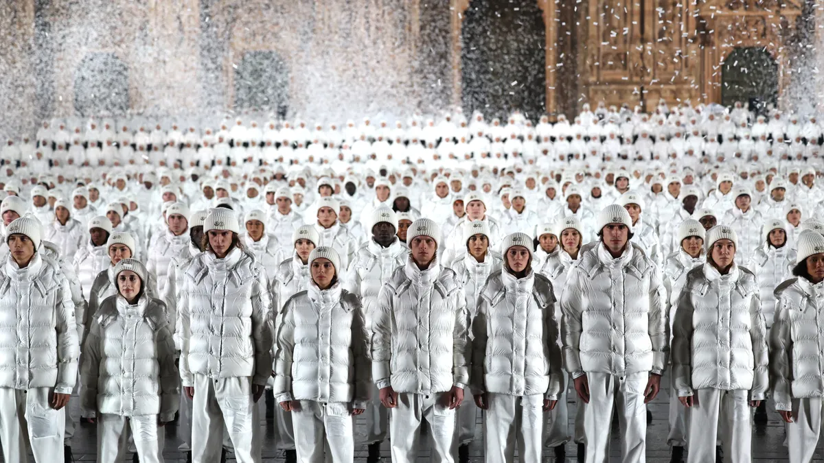 Dozens of models in white puffer jackets and hats line up in front a cathedral at nighttime for a Moncler fashion show.