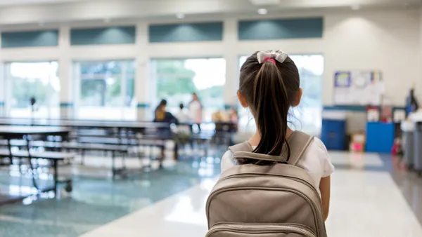 A student with a backpack has their back to the camera and is in a school lunchroom.