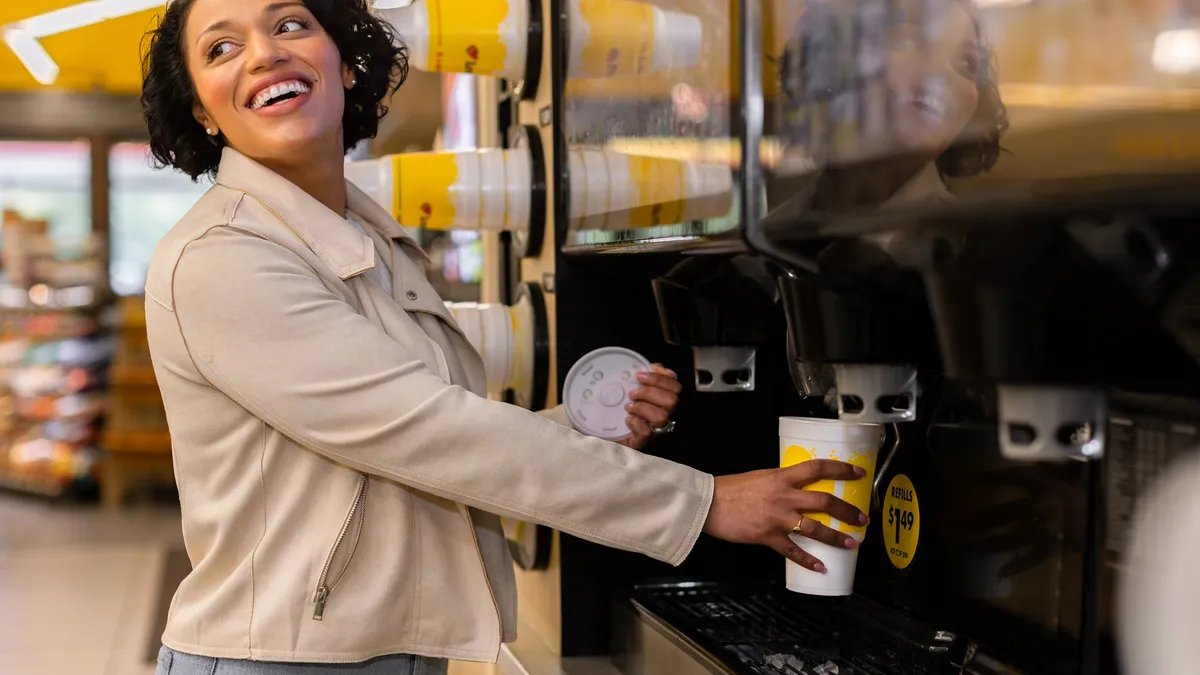 A photo of a person pouring a beverage from a large dispenser in a store into a white and yellow disposable cup. the person has a lid for the cup in the other hand.