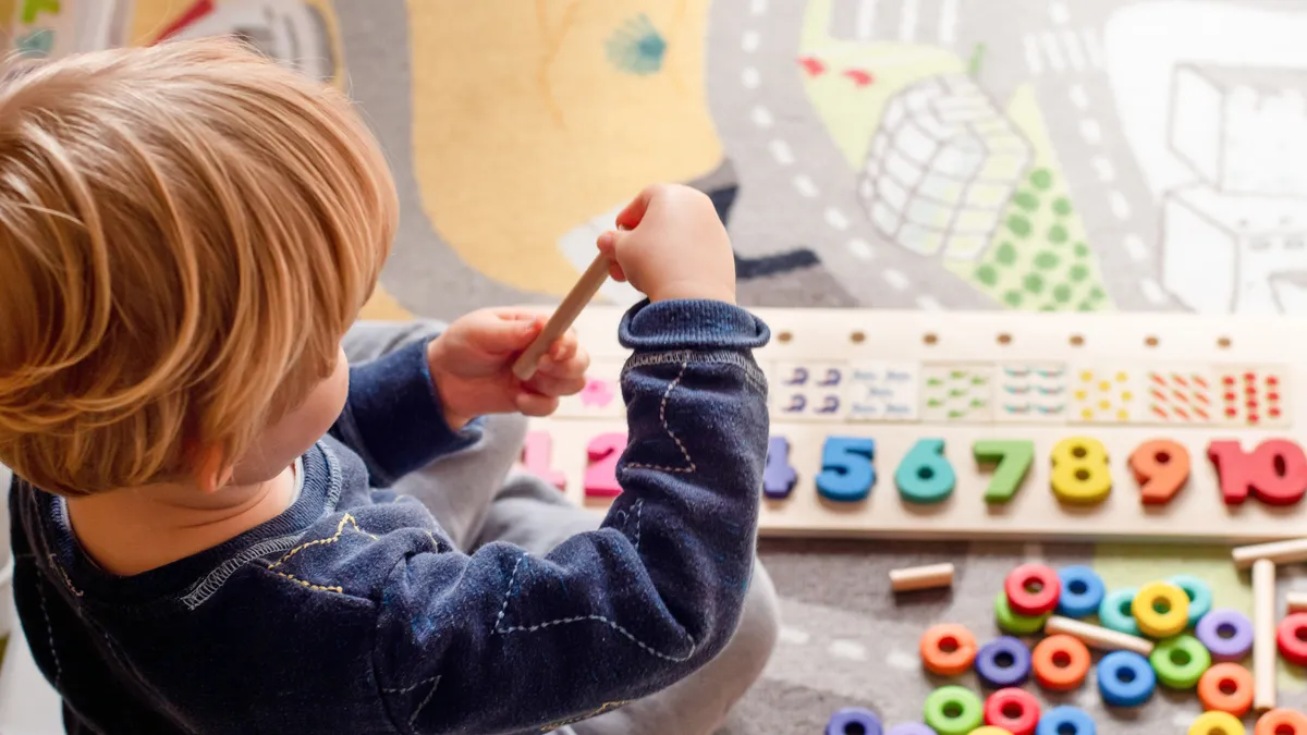 A bird's eye view of a toddler playing on the ground with colorful wooden numbers, wooden sticks and circles.