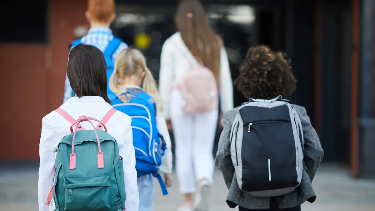 A group of students carrying backpacks are shown with their backs to the camera and walking toward a building.