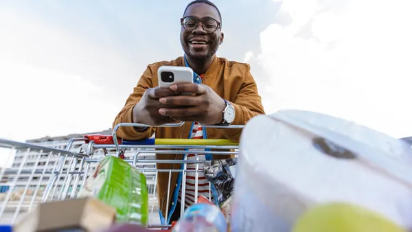 A smiling man looks at his phone while pushing a shopping cart filled with various groceries. The sky is bright, evoking a cheerful mood.