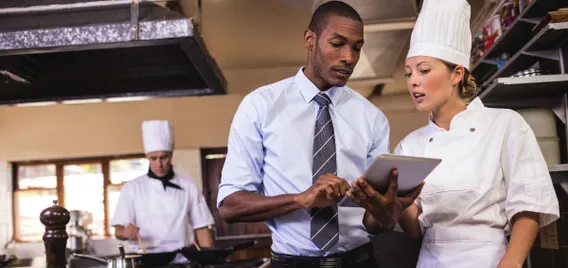 A man and a woman dressed as a chef look at a tablet.