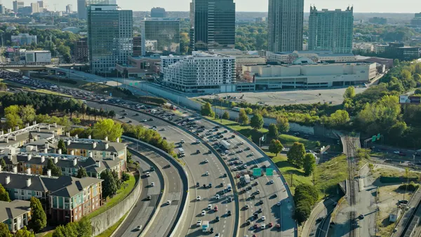 High-rise office towers and condominium buildings rise above a large shopping mall and parking structures in the mid-ground, while railroad tracks run parallel to the highway on the right side of the frame.
