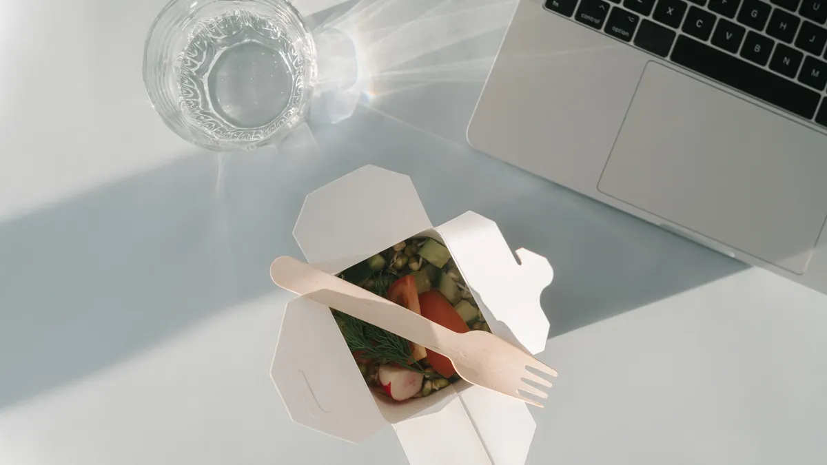 A glass, a laptop and some takeout with a compostable single-use spork