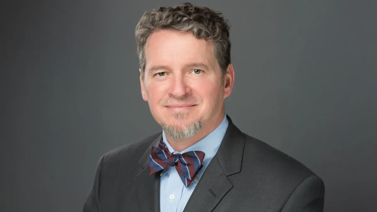 HCA Healthcare's Dr. Thomas Payne wears a gray suit jacket and blue and burgundy striped bow tie, against a gray background.