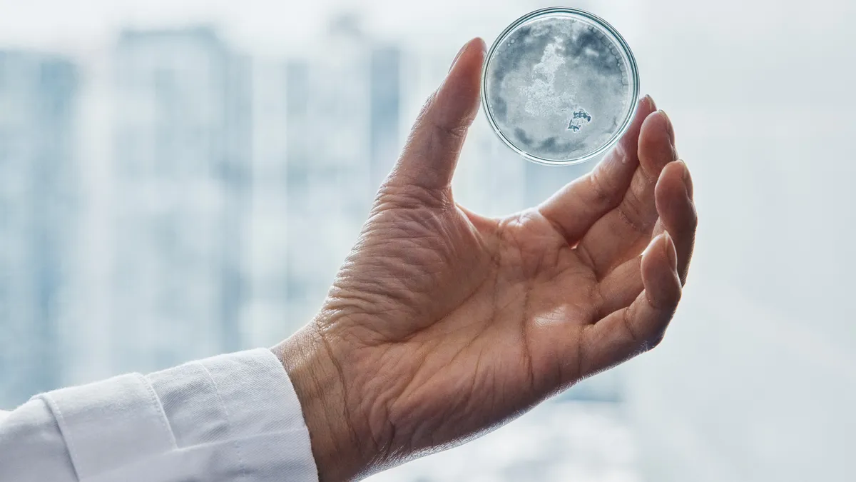 A hand in a white sleeve holds up a small sphere filled with water and microplastics against a blurred window.