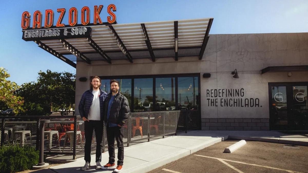 Two business owners standing in front of their restaurant named Gadzooks on a sunny day.