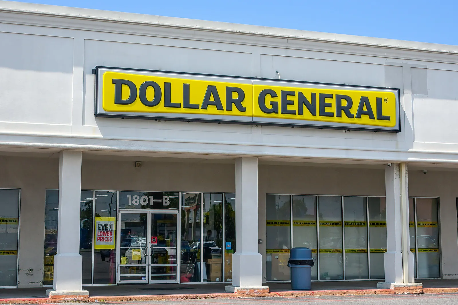 A plain white strip mall building with large glass windows. A bold yellow sign with black letters reads “DOLLAR GENERAL” above the entrance. The address “1801-B” is displayed on the door, and a trash bin is placed outside.