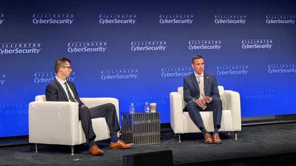 Two men sit on plush white chairs on a carpeted conference stage. Behind them, a backdrop displays a logo reading "Billington Cybersecurity."