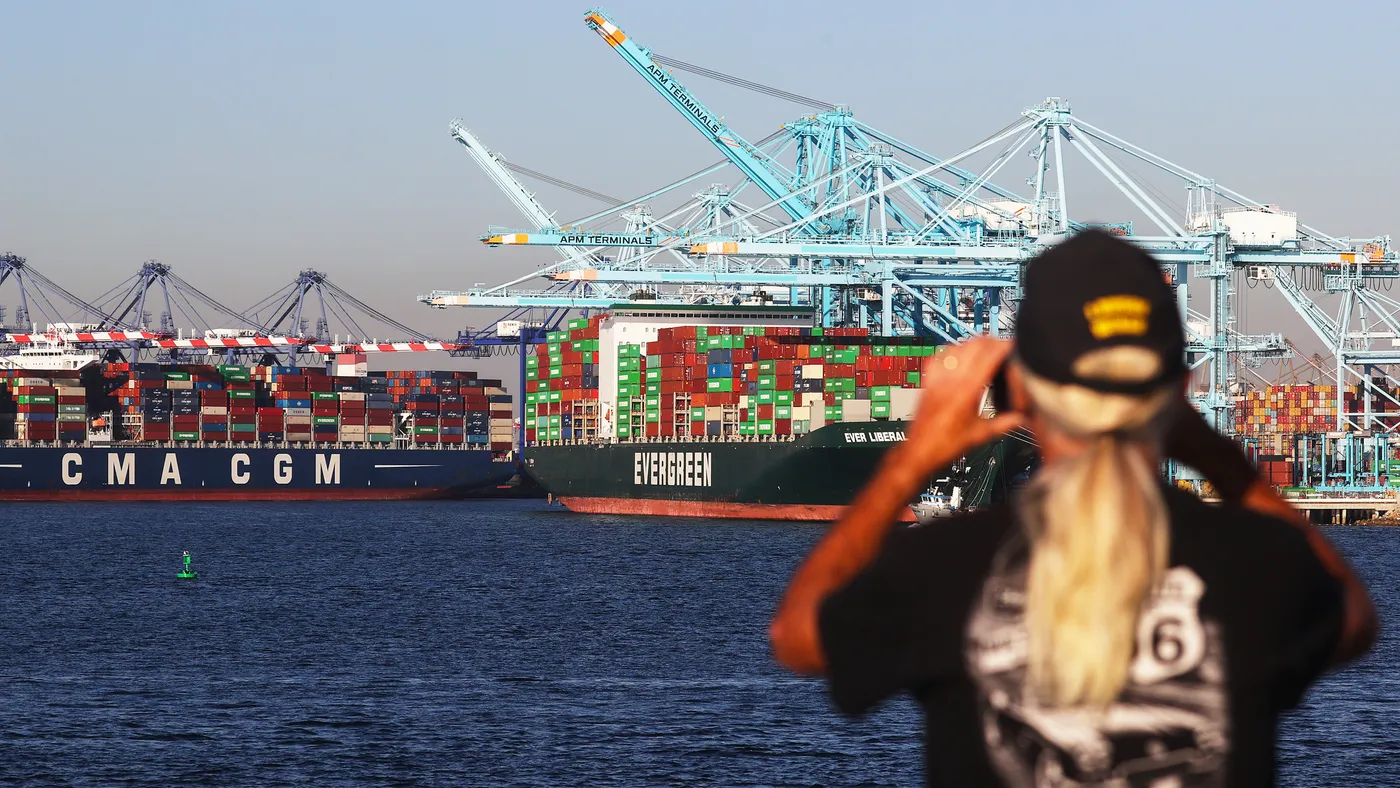 A person looks with binoculars at large cargo ships carrying containers.