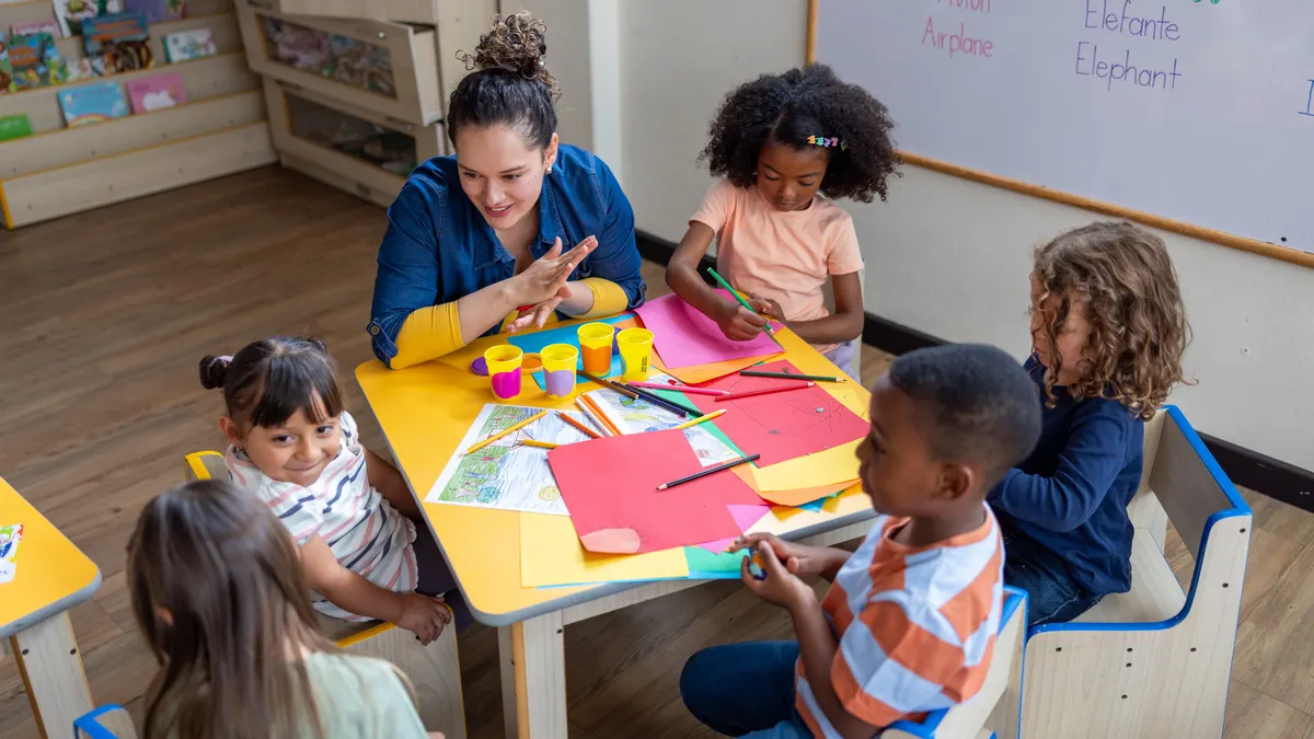 An adult sits at a small table with young students in a classroom. On the table is paper and Play-doh.