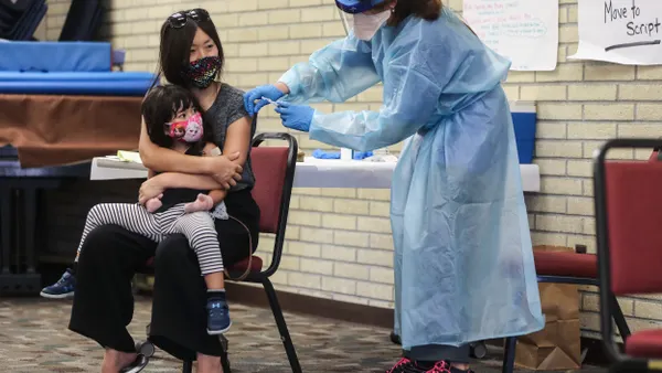 Person wearing mask sitting in chair with toddler in lap receives a shot from a person wearing mask and medical gown.