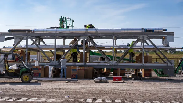A large piece of construction equipment looms in the foreground, on a bright, sunny day, made of steel piping.
