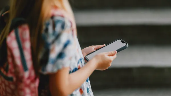 A student with a backpack is shown from the side from the shoulders down, In their hands is a cellphone