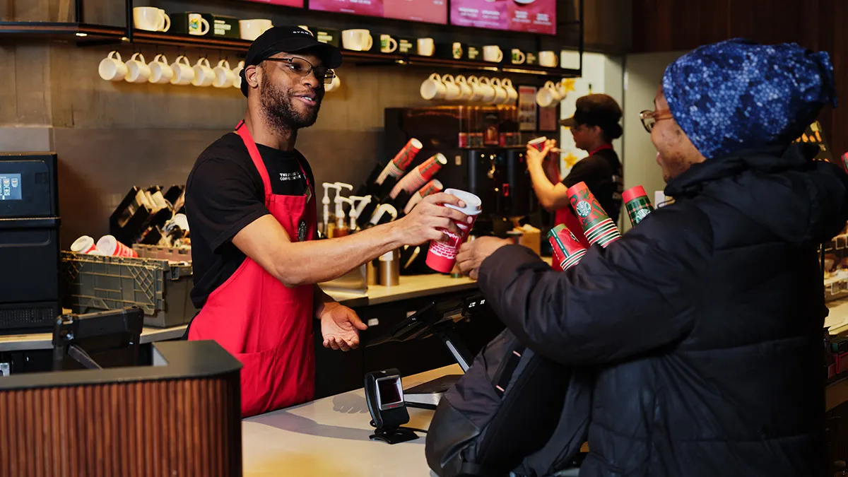 A person wearing a green apron hands off a reusable red cup to another person inside a coffee shop