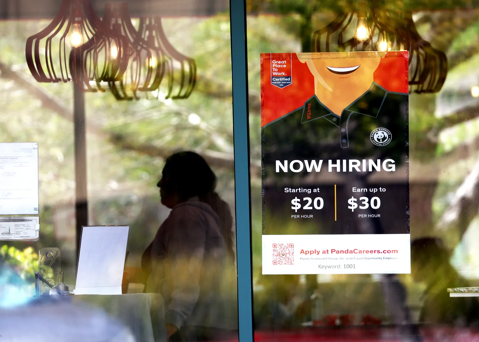 A "now hiring" sign is posted in a restaurant window, advertising an hourly pay rate of $20 to $30 per hour.