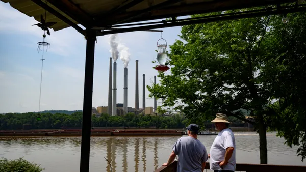 Two men stand near a river with power plant smokestacks in the background.