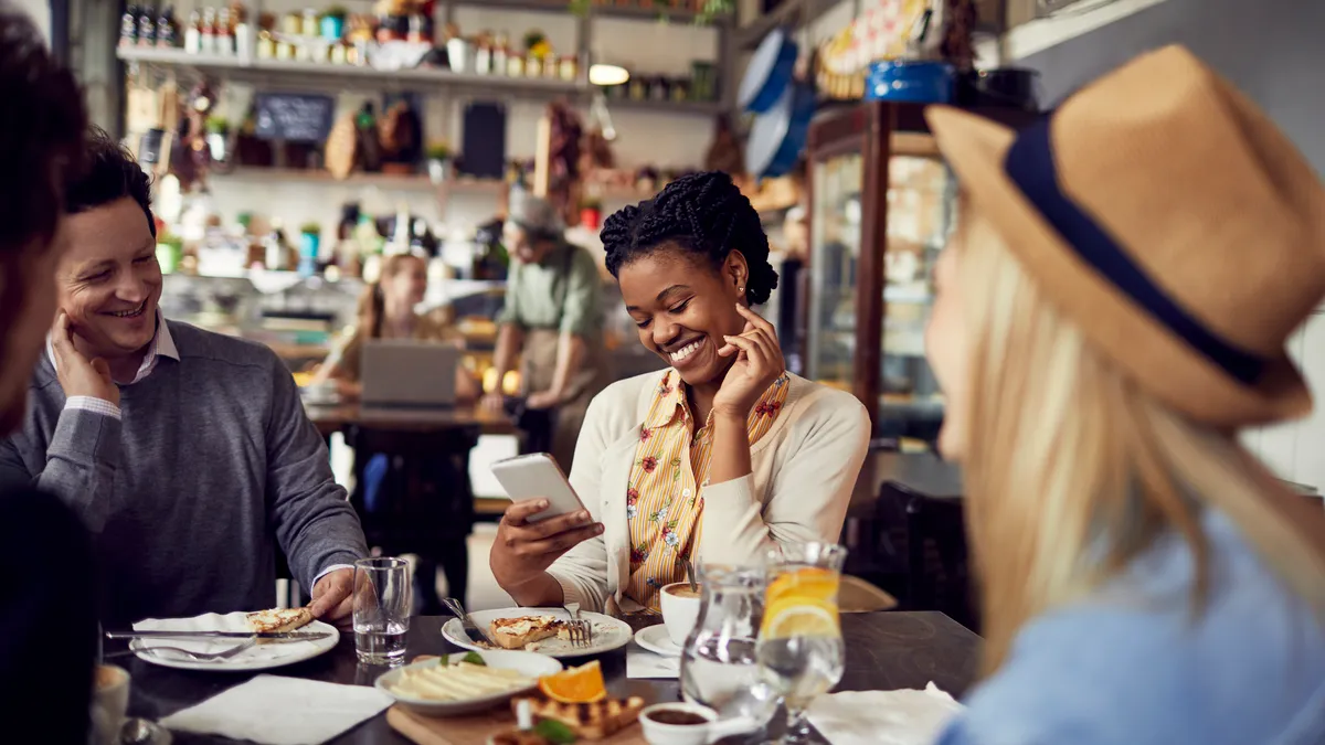 A group of people are enjoying their meal at a bright restaurant while one looks on a smartphone.