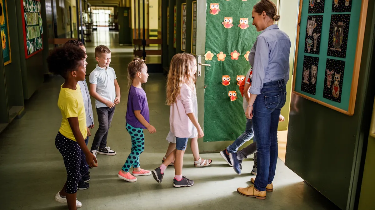 A line of students prepares to walk inside a classroom as they are greeted by a teacher.