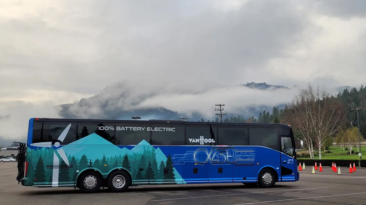 A large three-axle bus or motor coach, lettered as "100% battery electric," is parked in a parking lot under a cloudy sky.