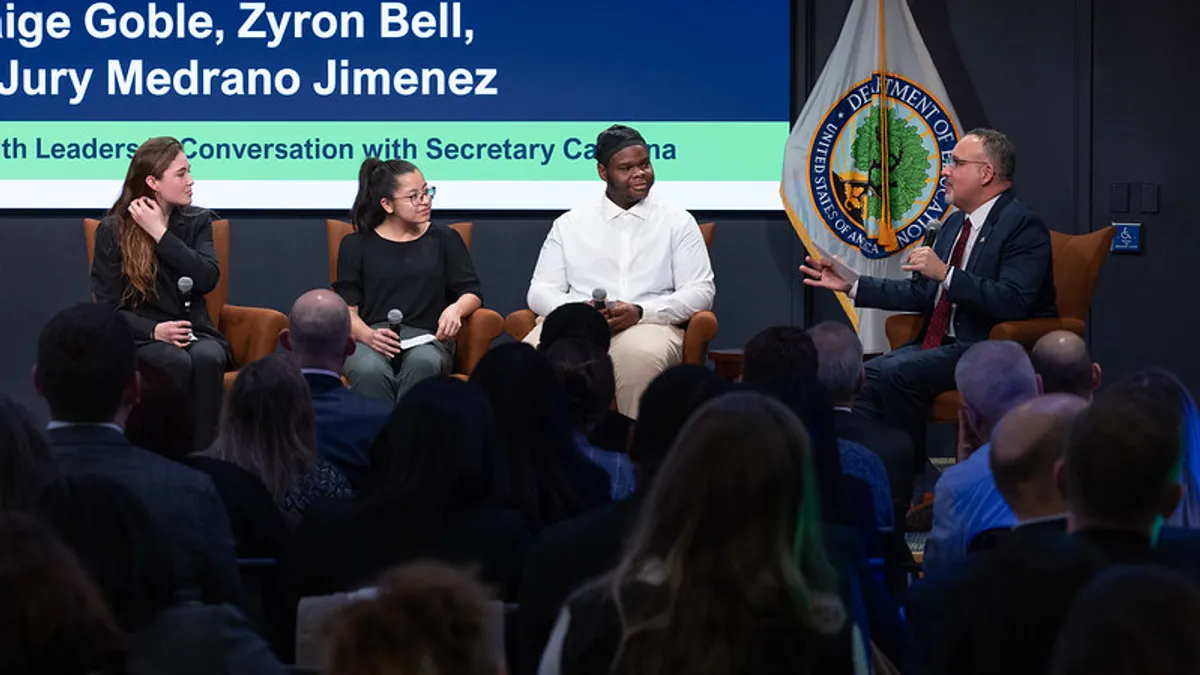 Four people are sitting on chairs on a stage. One person has a microphone. There is a screen and a U.S. flag behind the people.