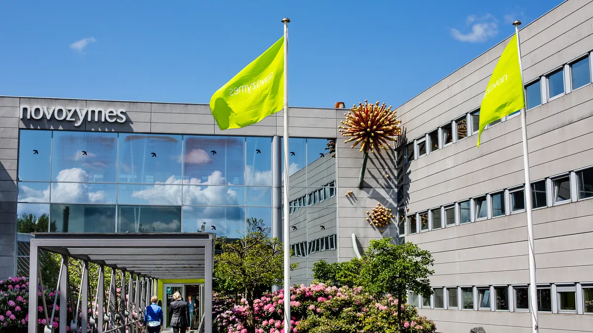 Bright green flags fly in front of the L-shaped Novozymes office building, with a mirrored wall straight ahead and white brick with small windows to the right. People walk on a covered path.