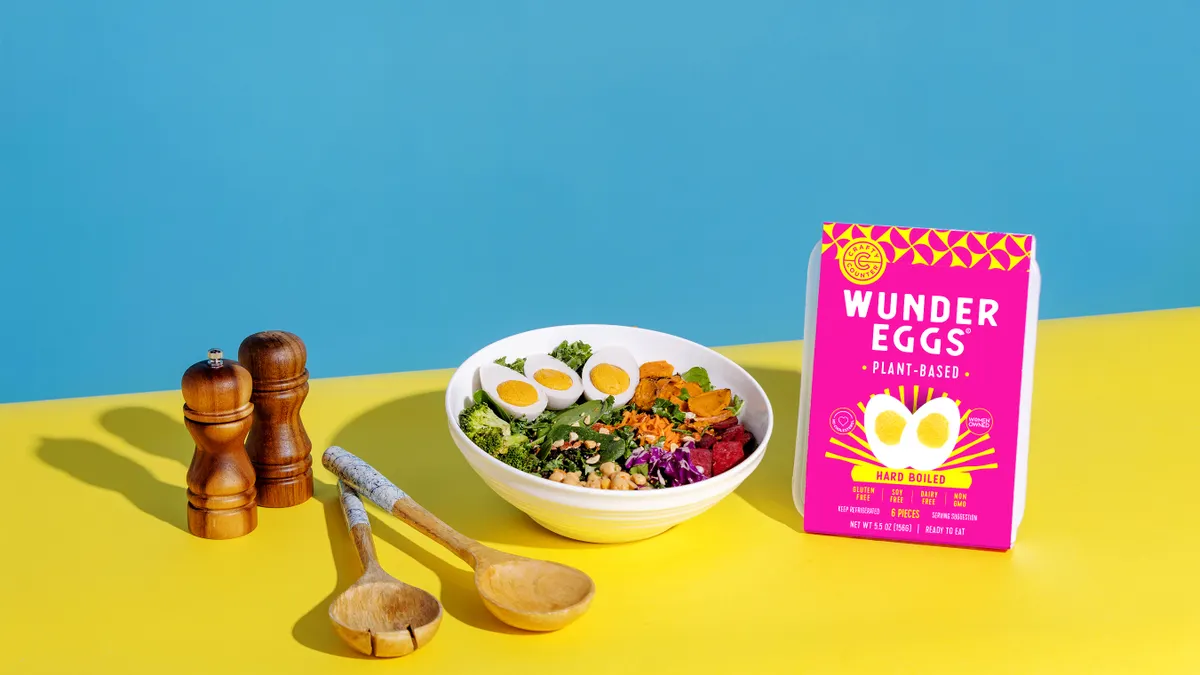 A white bowl with a salad that has three WunderEgg halves sits on a yellow surface. A pink-labeled package of WunderEggs is at right, and wooden salad tongs and salt and pepper shakers are at left