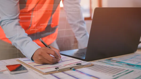 A person wearing a long-sleeved shirt and an orange vest holds a pencil above a long white piece of paper on a desk.