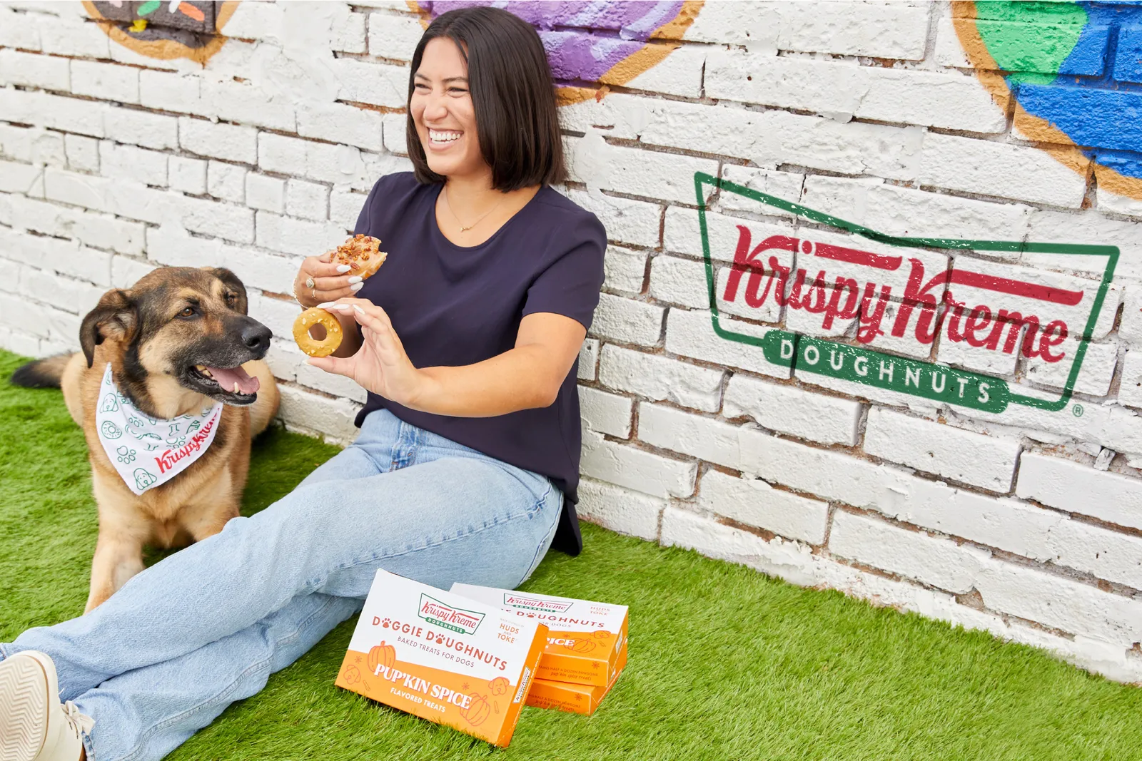 A woman sits in the grass with a dog and holds up a donut and a dog biscuit.